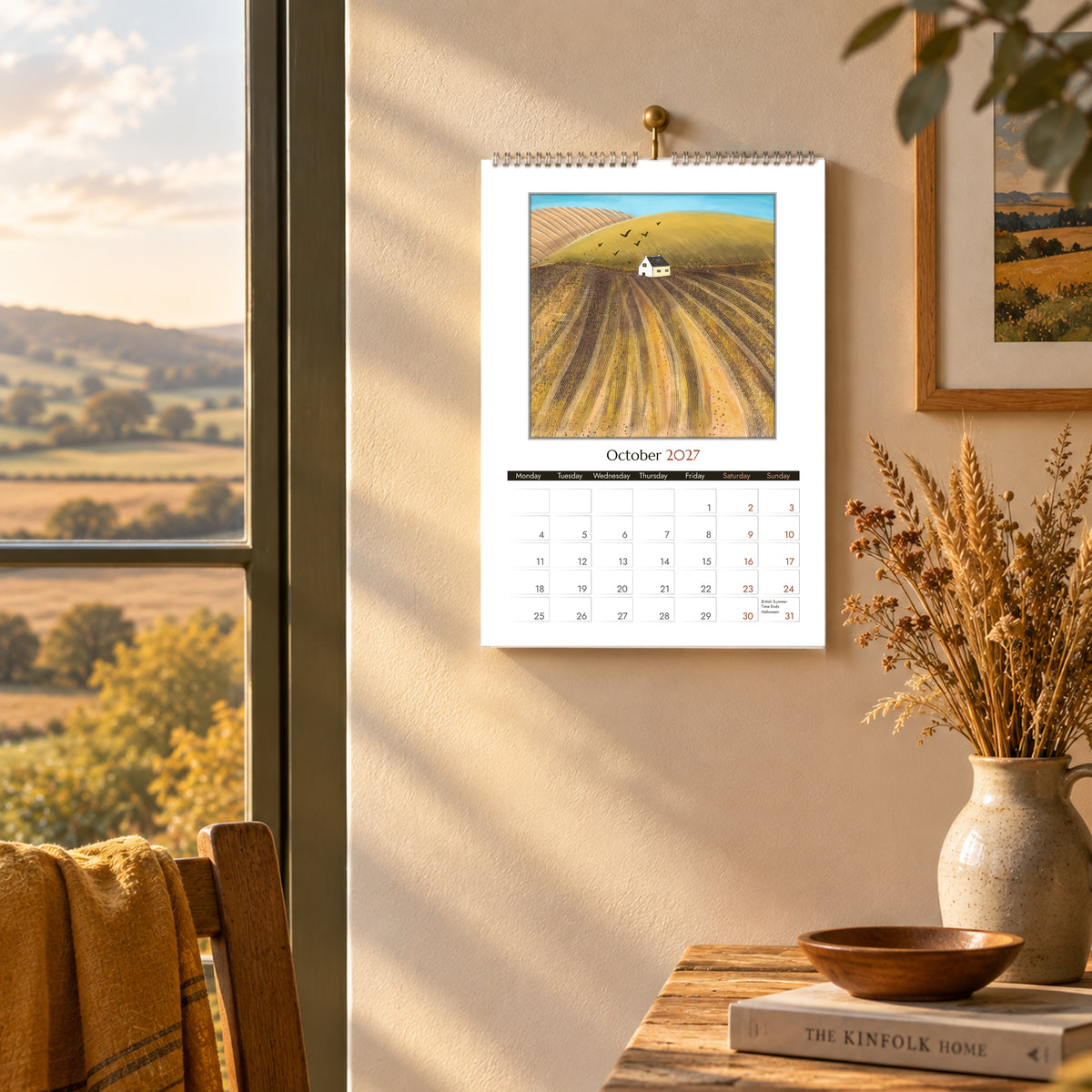 October 2027 calendar page featuring golden autumn fields and a white cottage, hanging in a warm rustic interior.