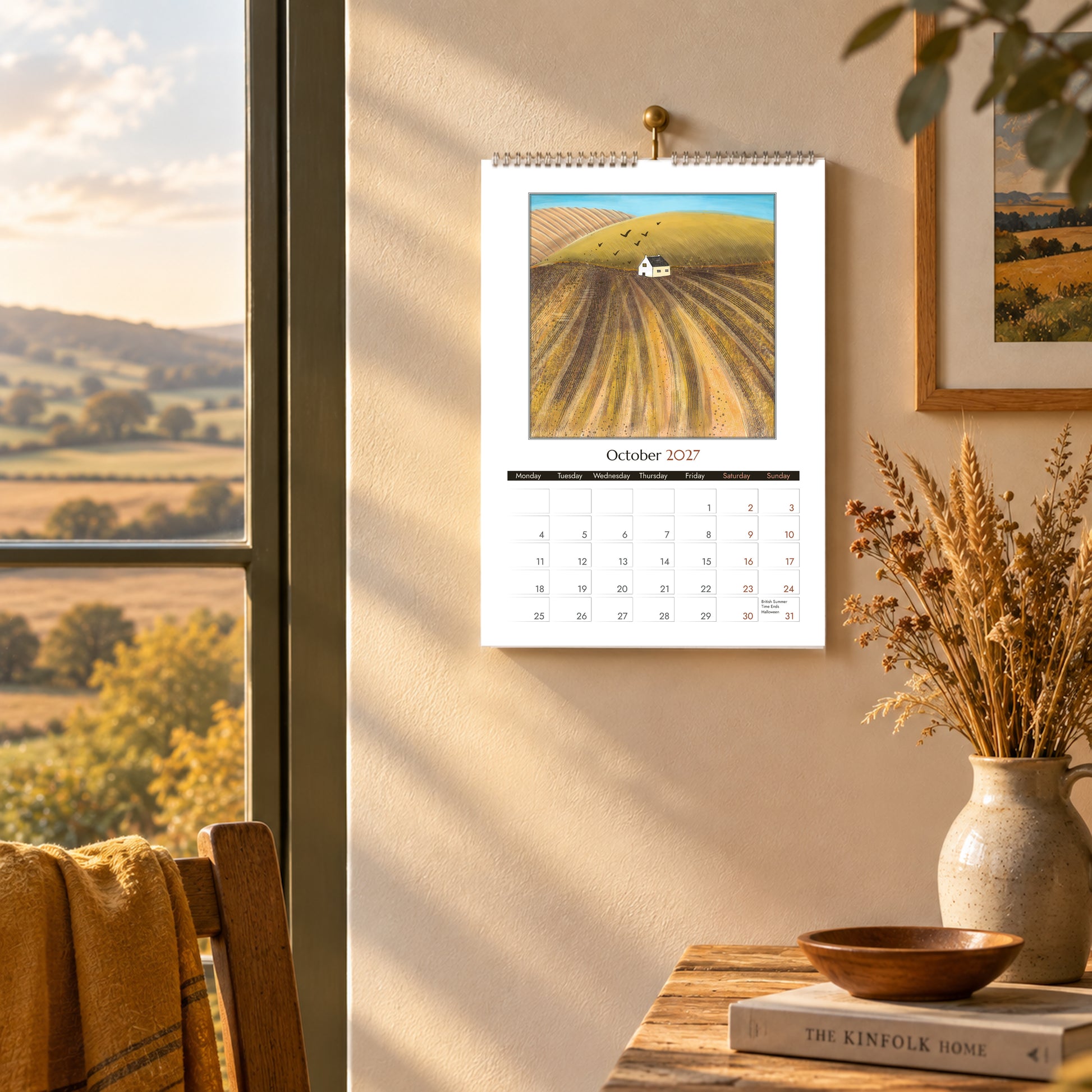 October 2027 calendar page featuring golden autumn fields and a white cottage, hanging in a warm rustic interior.