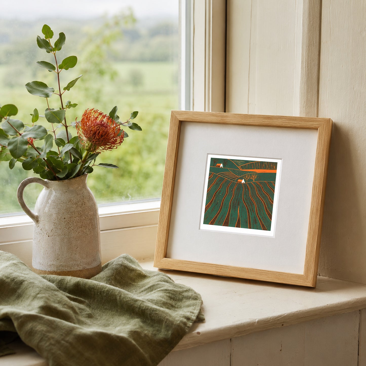 Mini linocut print framed in pale oak, propped on a windowsill with a rose in a stone jug