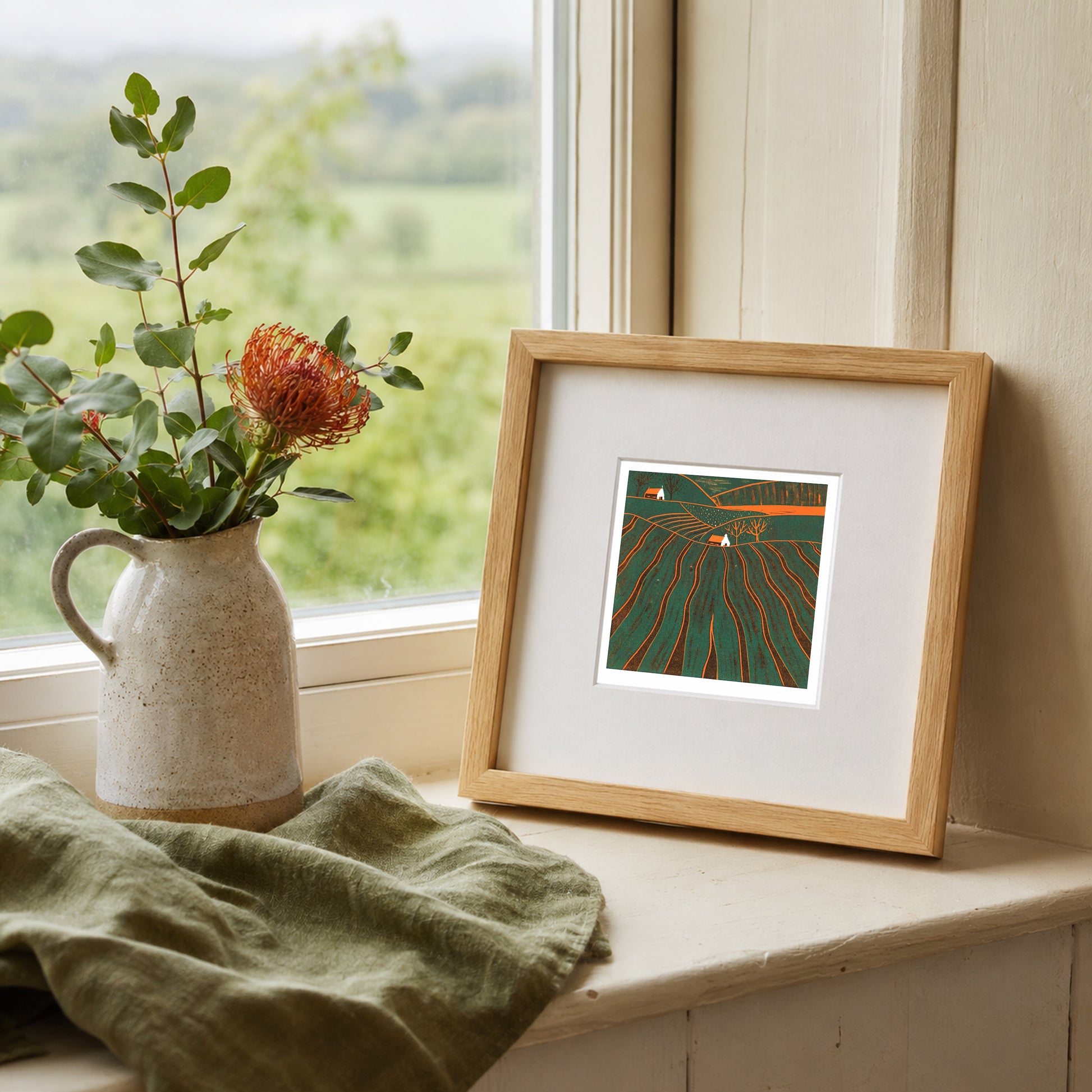 Mini linocut print framed in pale oak, propped on a windowsill with a rose in a stone jug