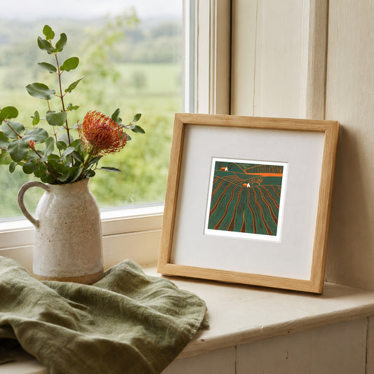 Mini linocut print framed in pale oak, propped on a windowsill with a rose in a stone jug