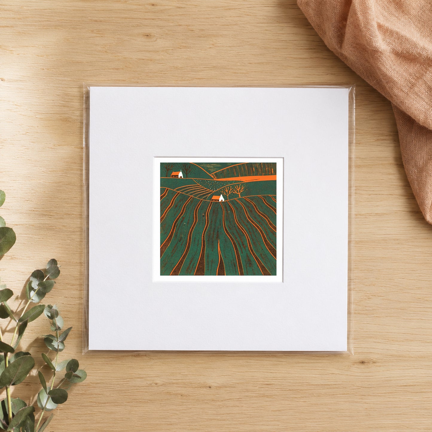 Mini linocut furrowed hills print in a white window mount, photographed flat on warm oak with eucalyptus