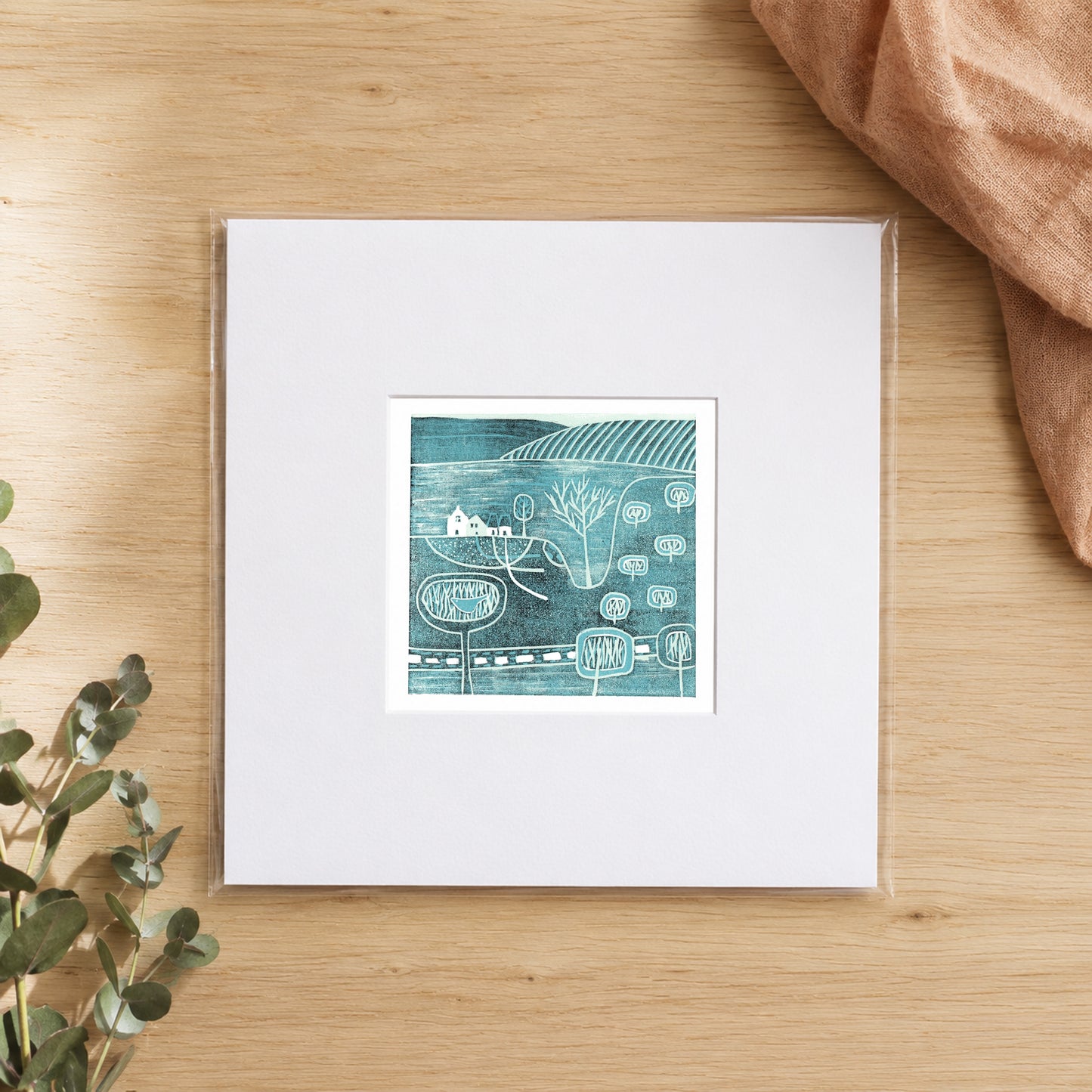 Mini linocut teal village print in a white window mount, photographed flat on warm oak with eucalyptus
