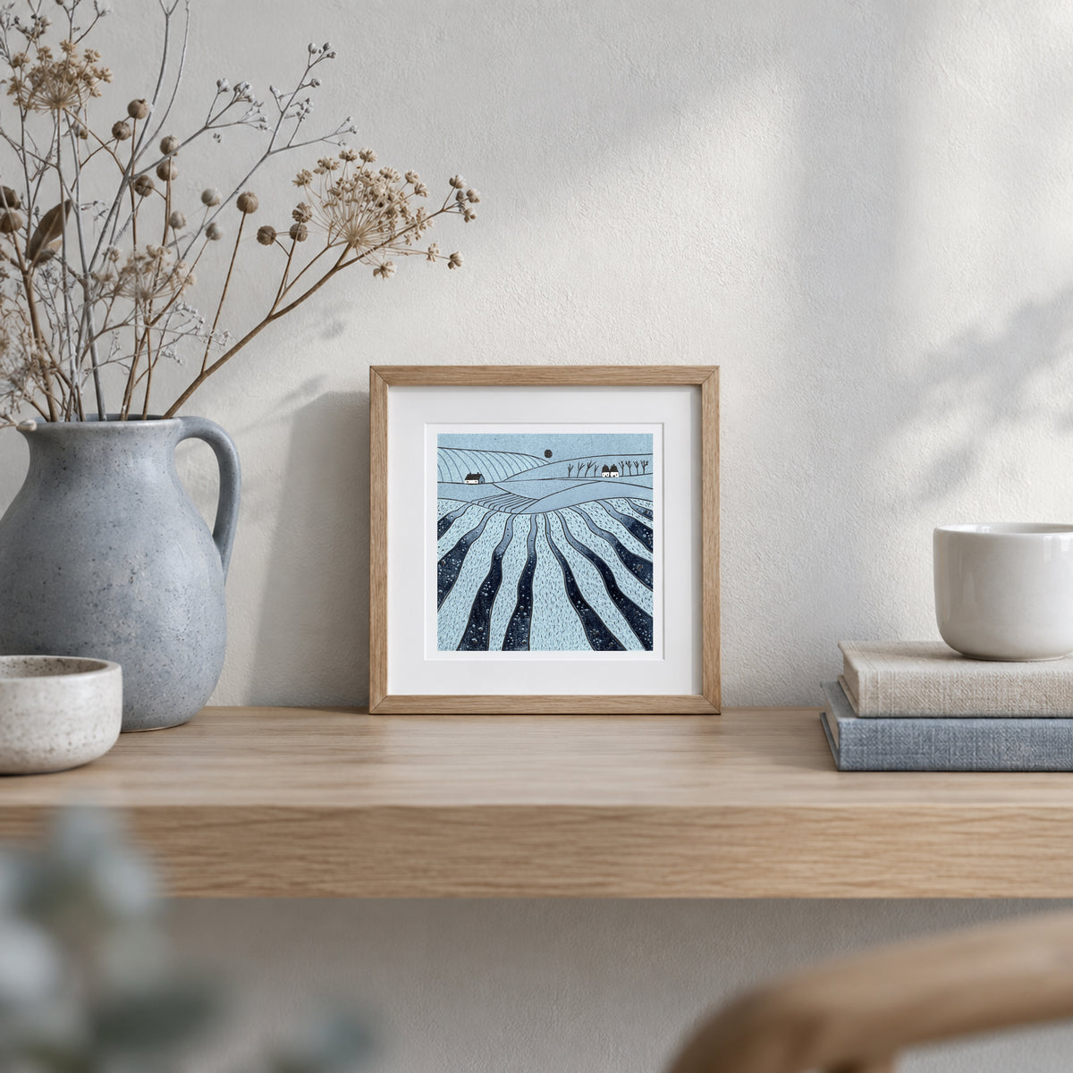 Where the Hills Breathe blue landscape linocut displayed in a natural wood frame on a shelf beside a ceramic vase of dried flowers.