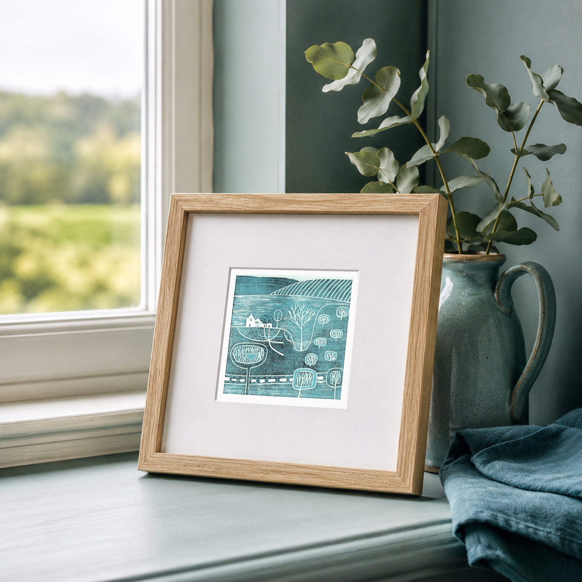 Mini linocut print framed in pale oak, propped on a windowsill with eucalyptus in a stone jug