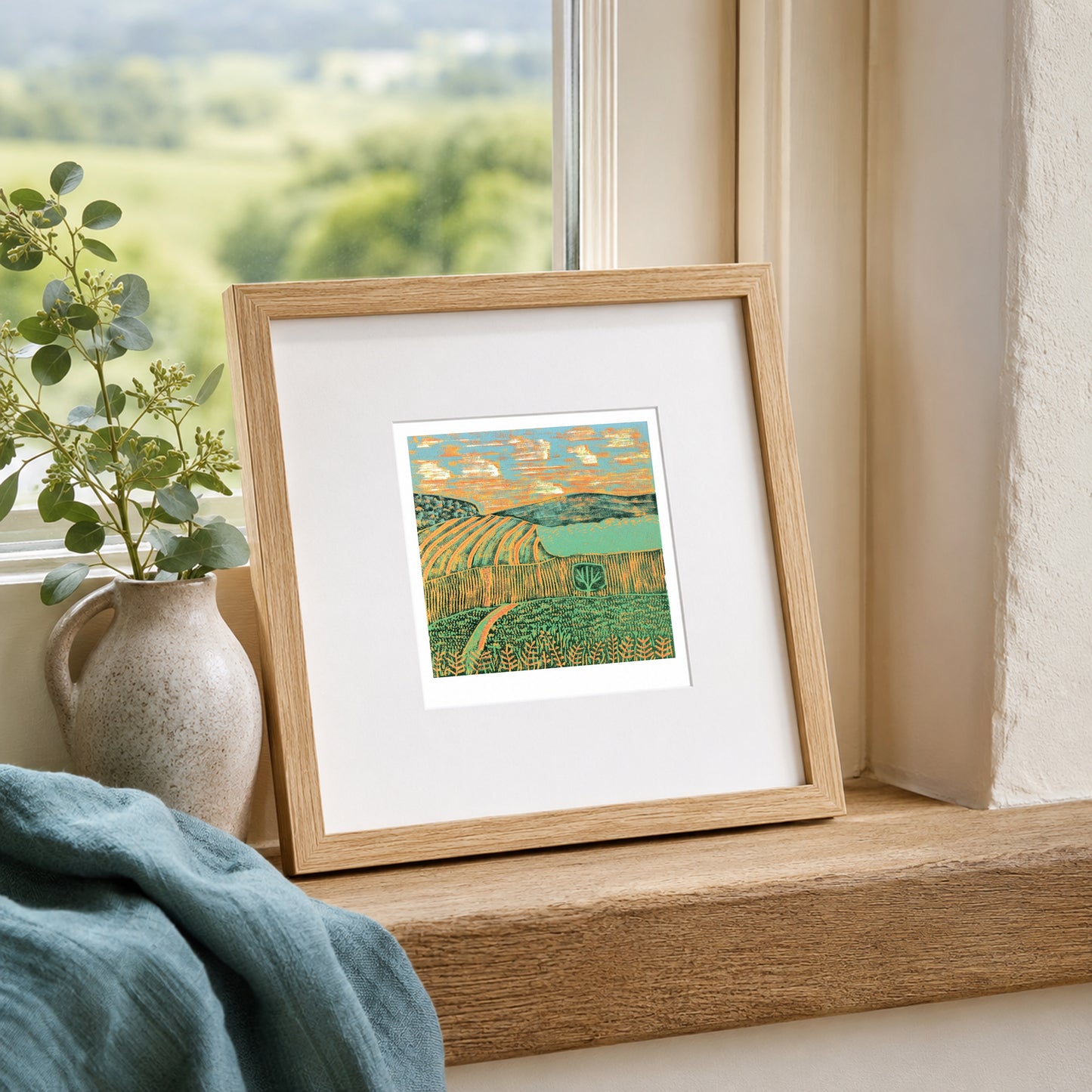 Mini linocut print framed in pale oak, propped on a windowsill with a vase and soft blue throw