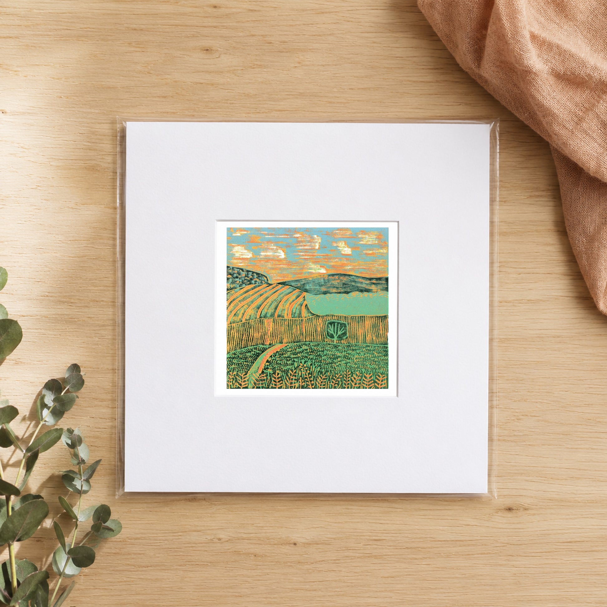 Mini linocut fields print in a white window mount, photographed flat on warm oak with eucalyptus