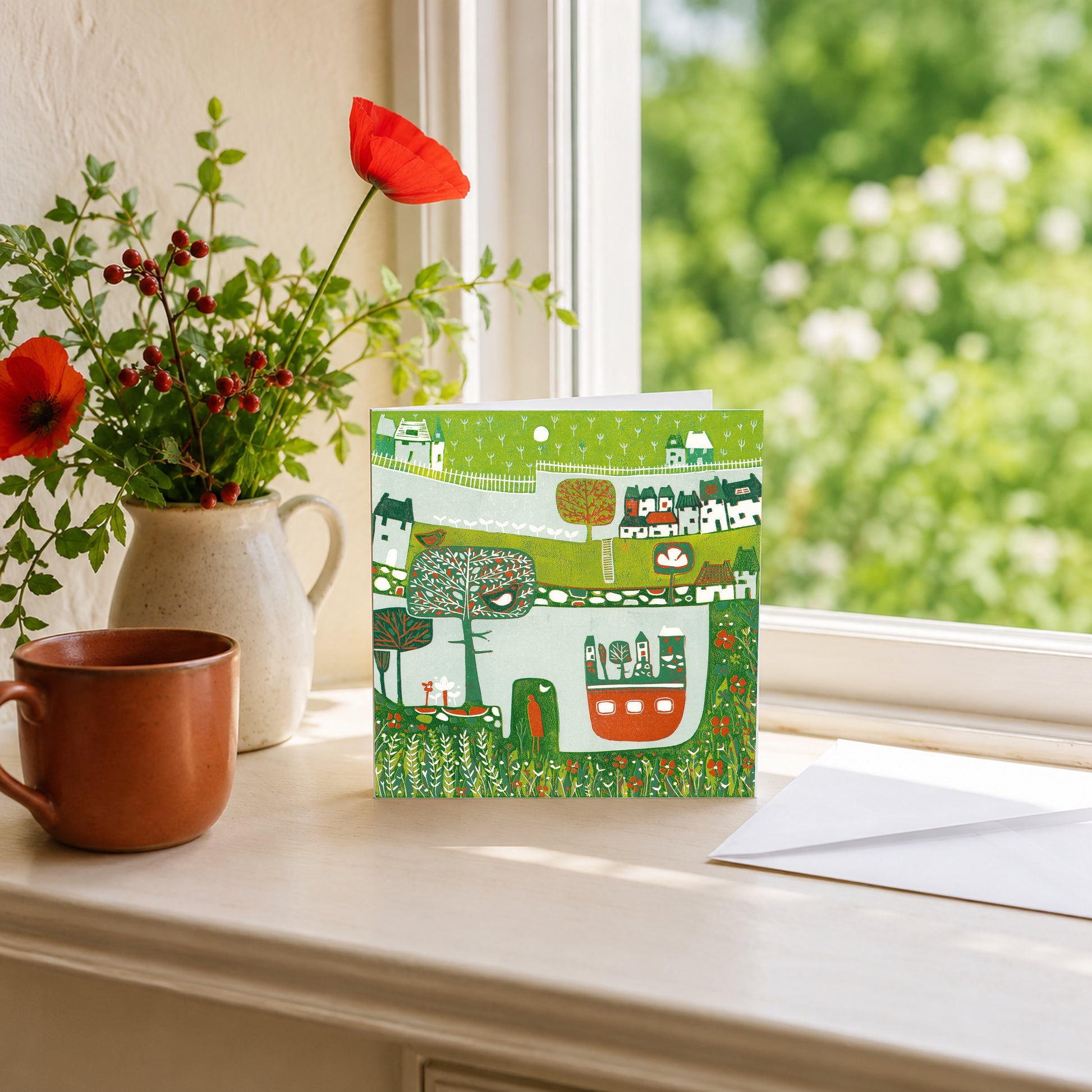 Linocut green landscape greeting card propped on a windowsill with a stone jug of red flowers