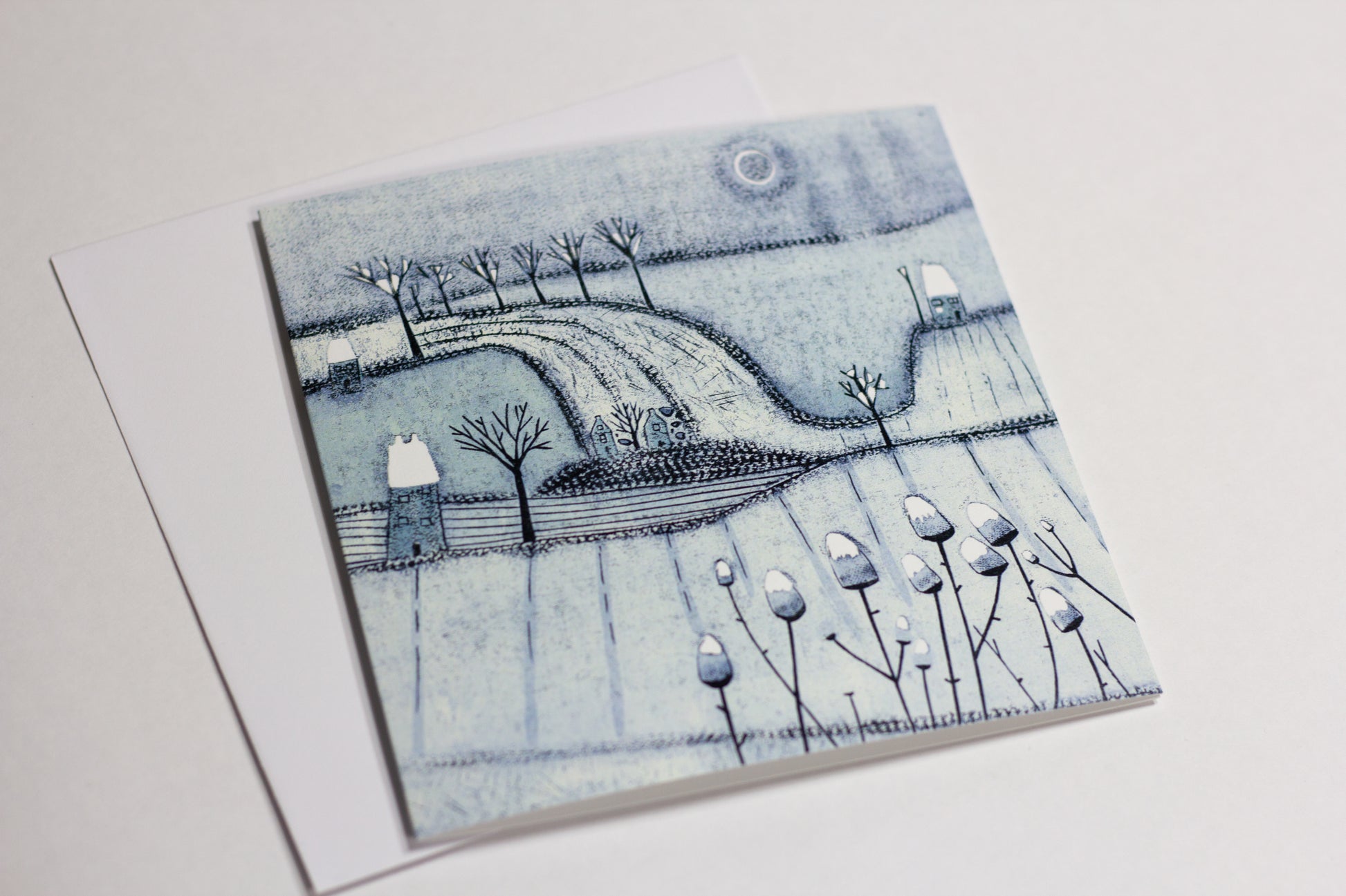 Close-up of a blue-grey winter Christmas card featuring trees, snowy fields and cottages.