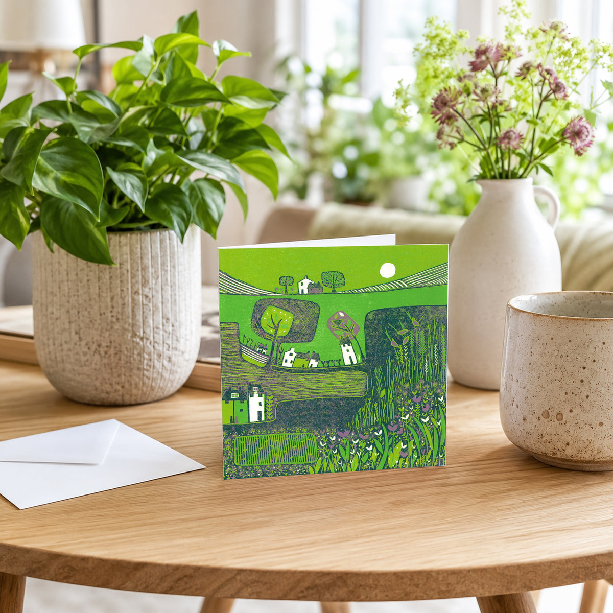 Whispering Melodies green linocut greeting card displayed on a wooden table beside a potted plant and a vase of flowers.