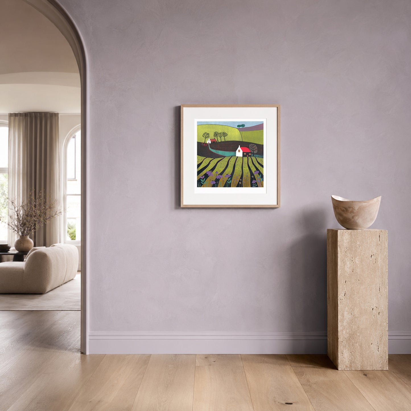 Linocut landscape print in an oak frame on a soft lilac plaster wall beside a travertine plinth.