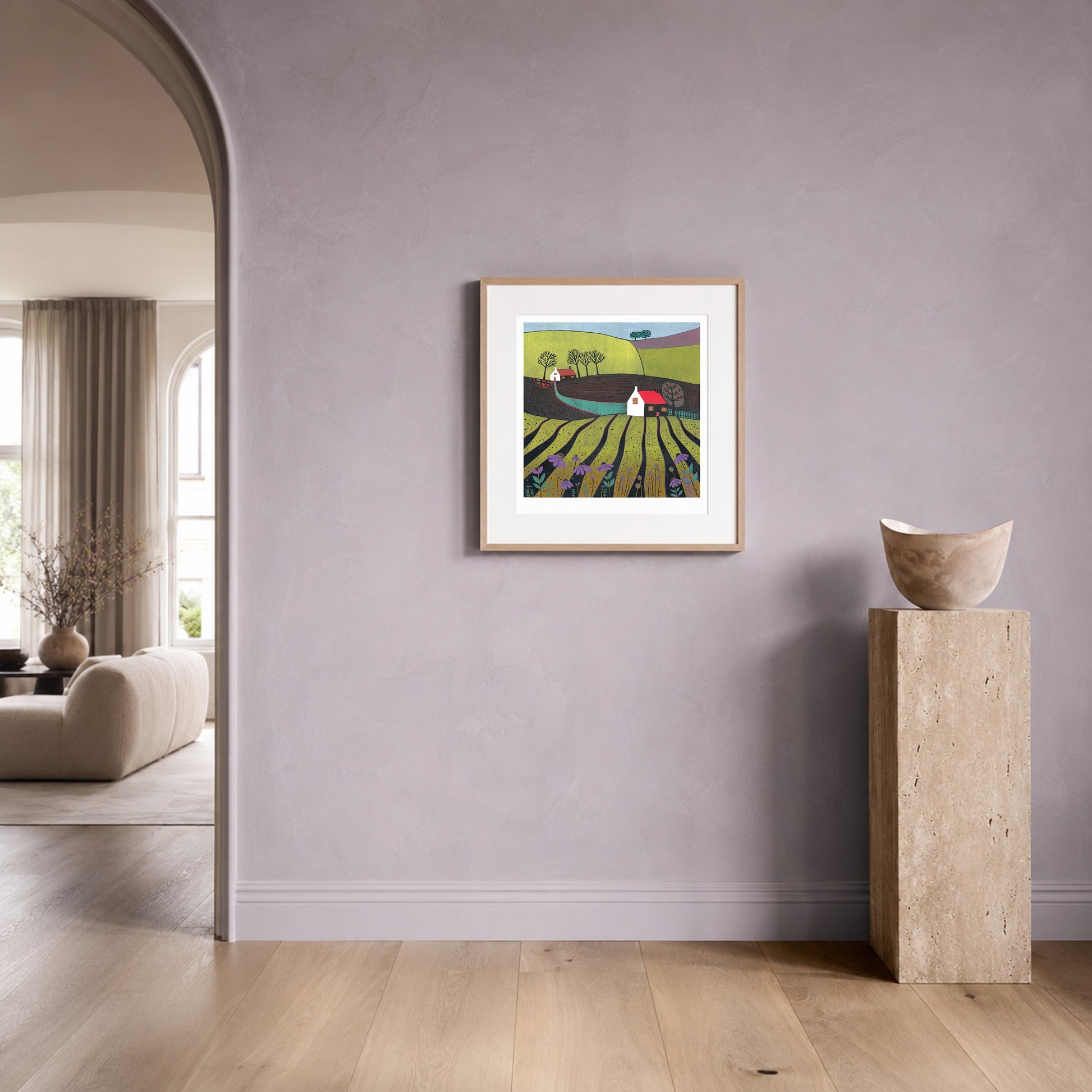Linocut landscape print in an oak frame on a soft lilac plaster wall beside a travertine plinth.