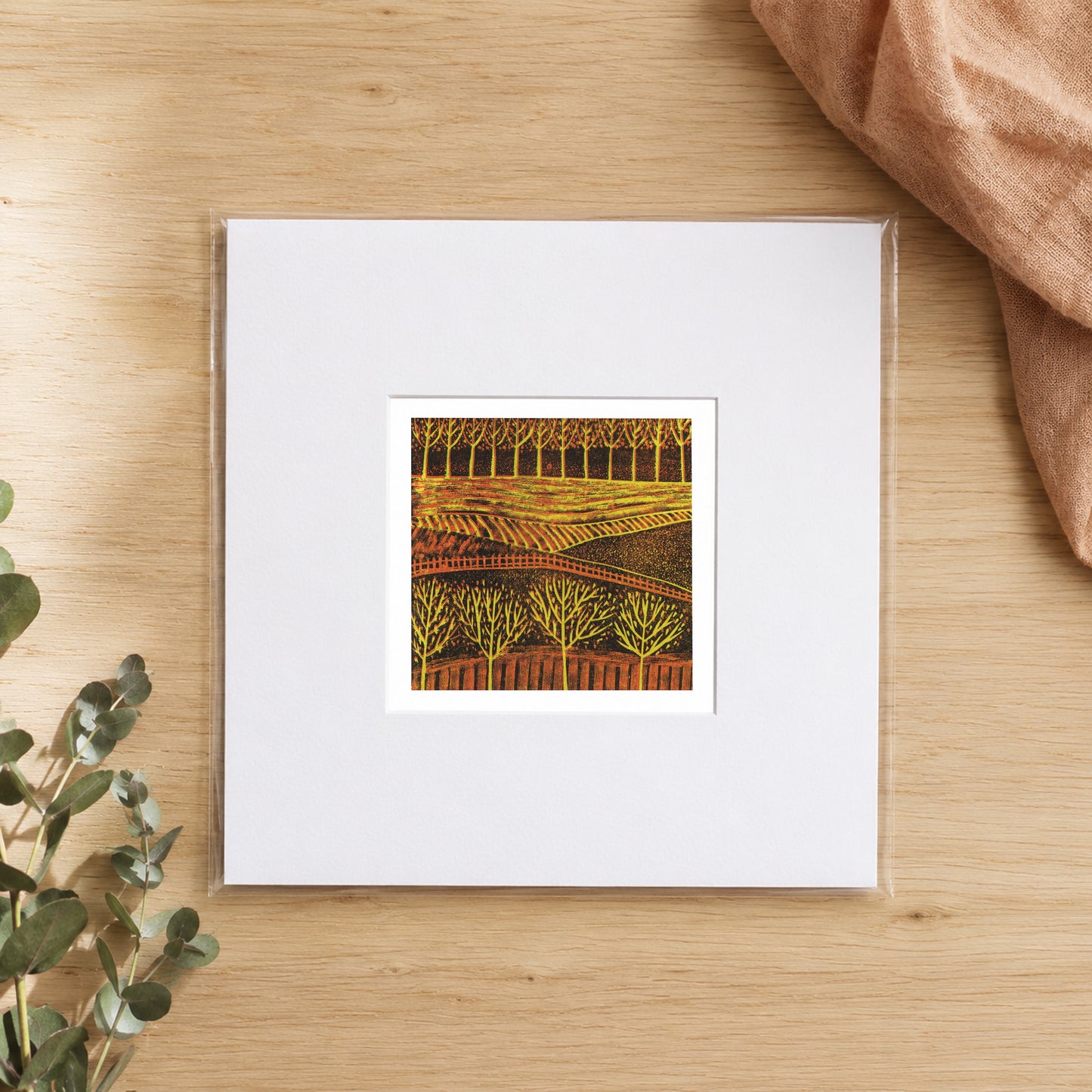 Mini linocut autumn trees print in a white window mount, photographed flat on warm oak with eucalyptus
