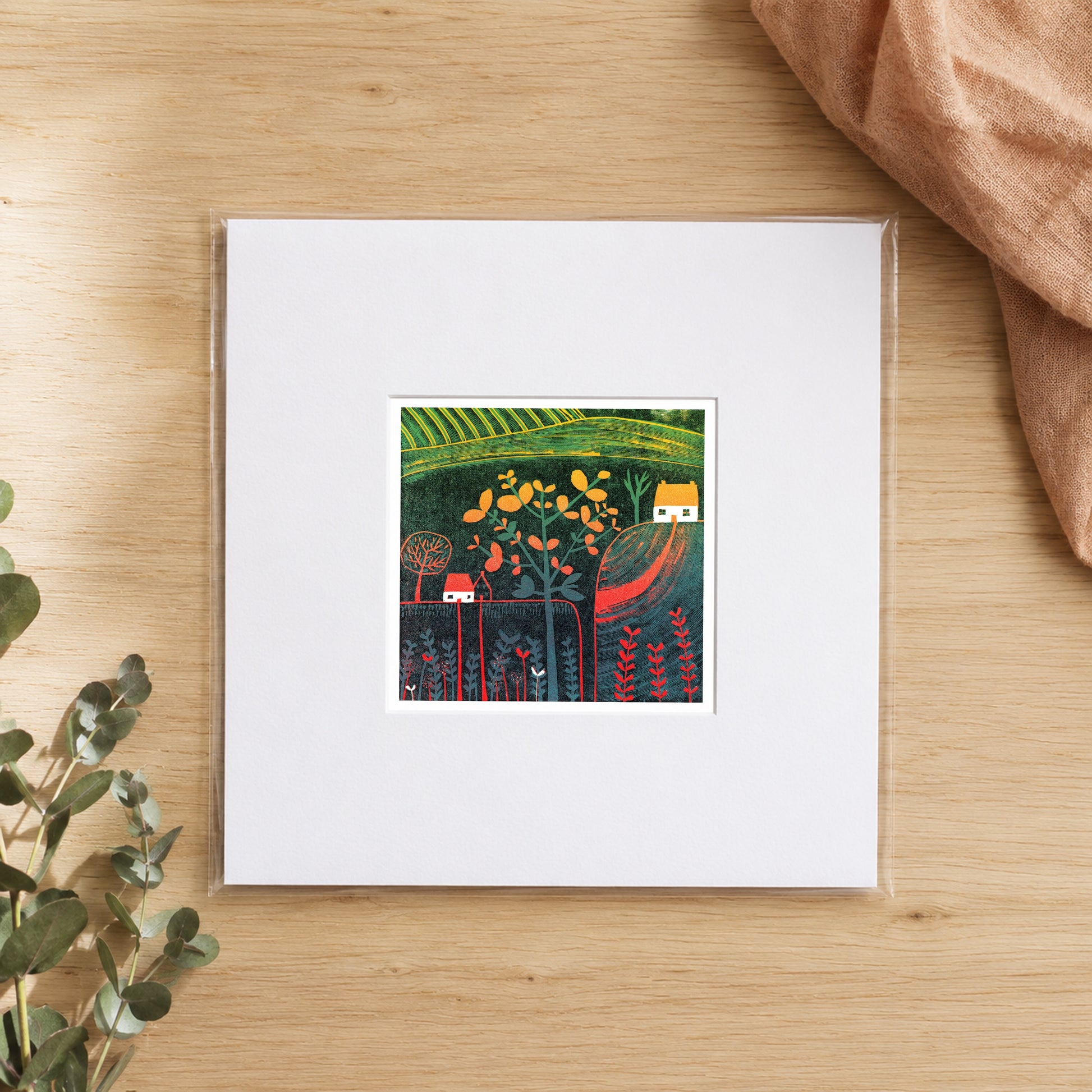 Mini linocut autumn cottage print in a white window mount, photographed flat on warm oak with eucalyptus