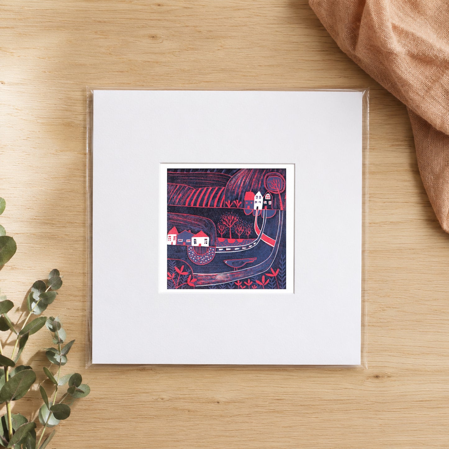 Mini linocut midnight village print in a white window mount, photographed flat on warm oak with eucalyptus