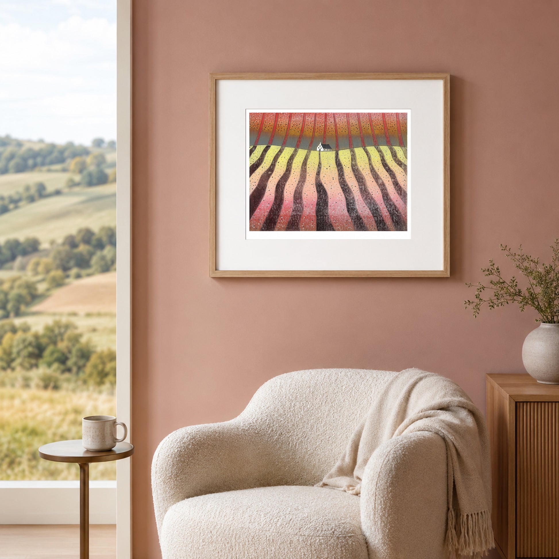 Framed print on a soft blush wall beside a window showing rolling green countryside, above a pale armchair.
