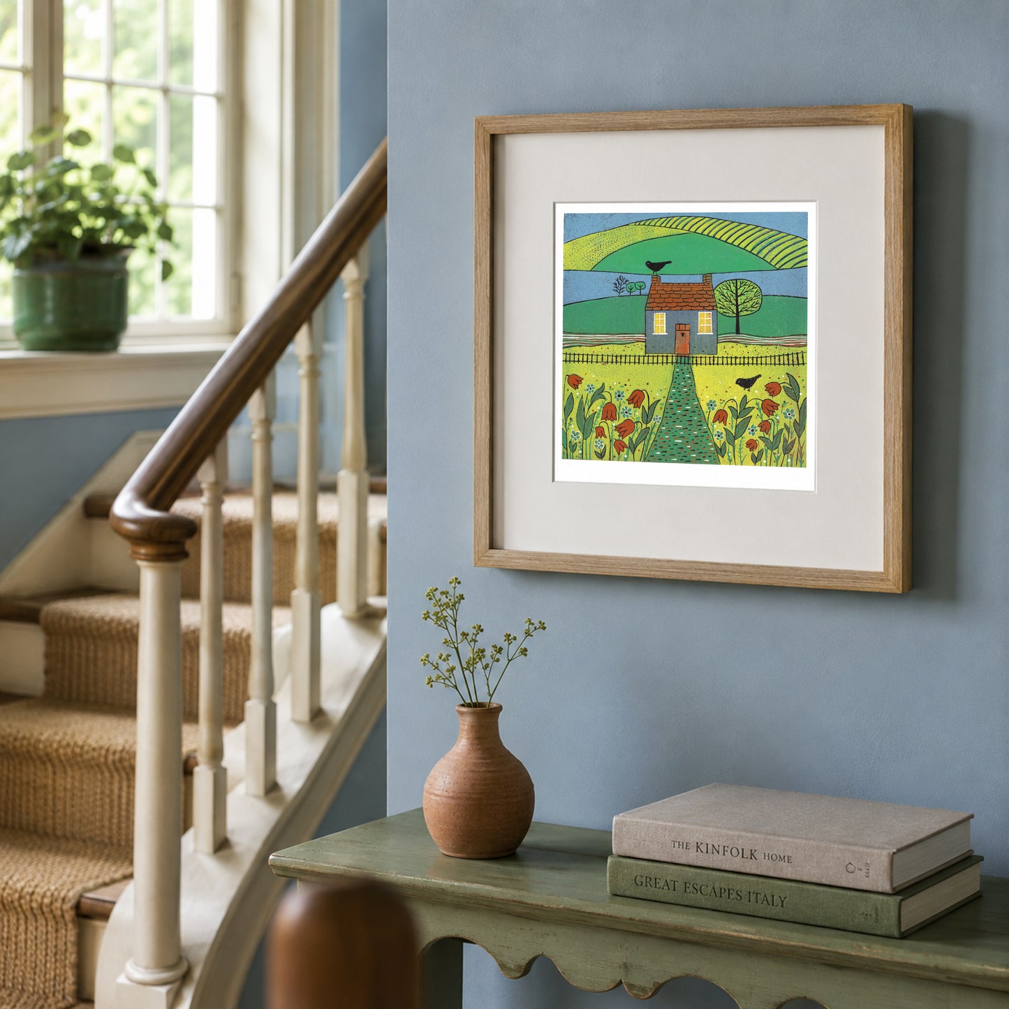 Framed Beyond the Garden linocut print with a cottage, birds and green hills displayed on a blue hallway wall beside a staircase.