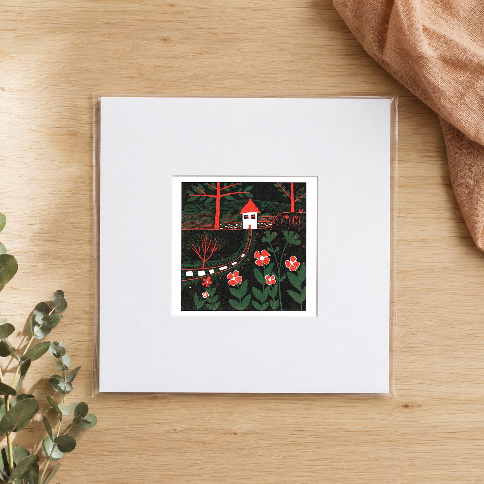 Mini linocut red house print in a white window mount, photographed flat on warm oak with eucalyptus