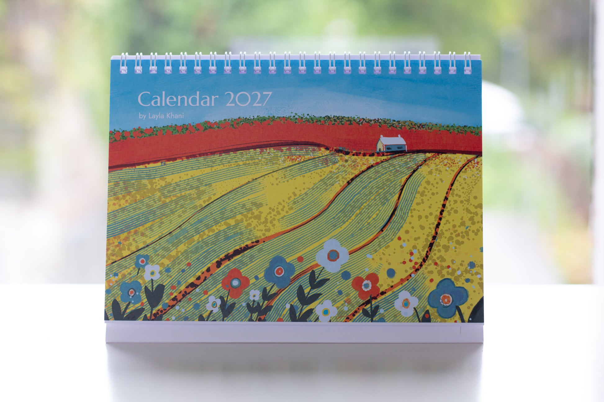 Photograph of the printed 2027 desk calendar cover showing yellow fields, flowers, white spiral binding and freestanding card stand.