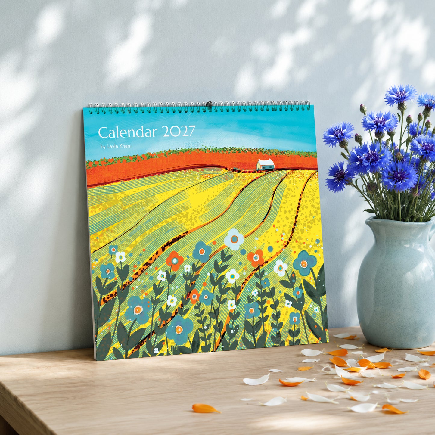 2027 large square art calendar with colourful countryside painting, displayed on a wooden table