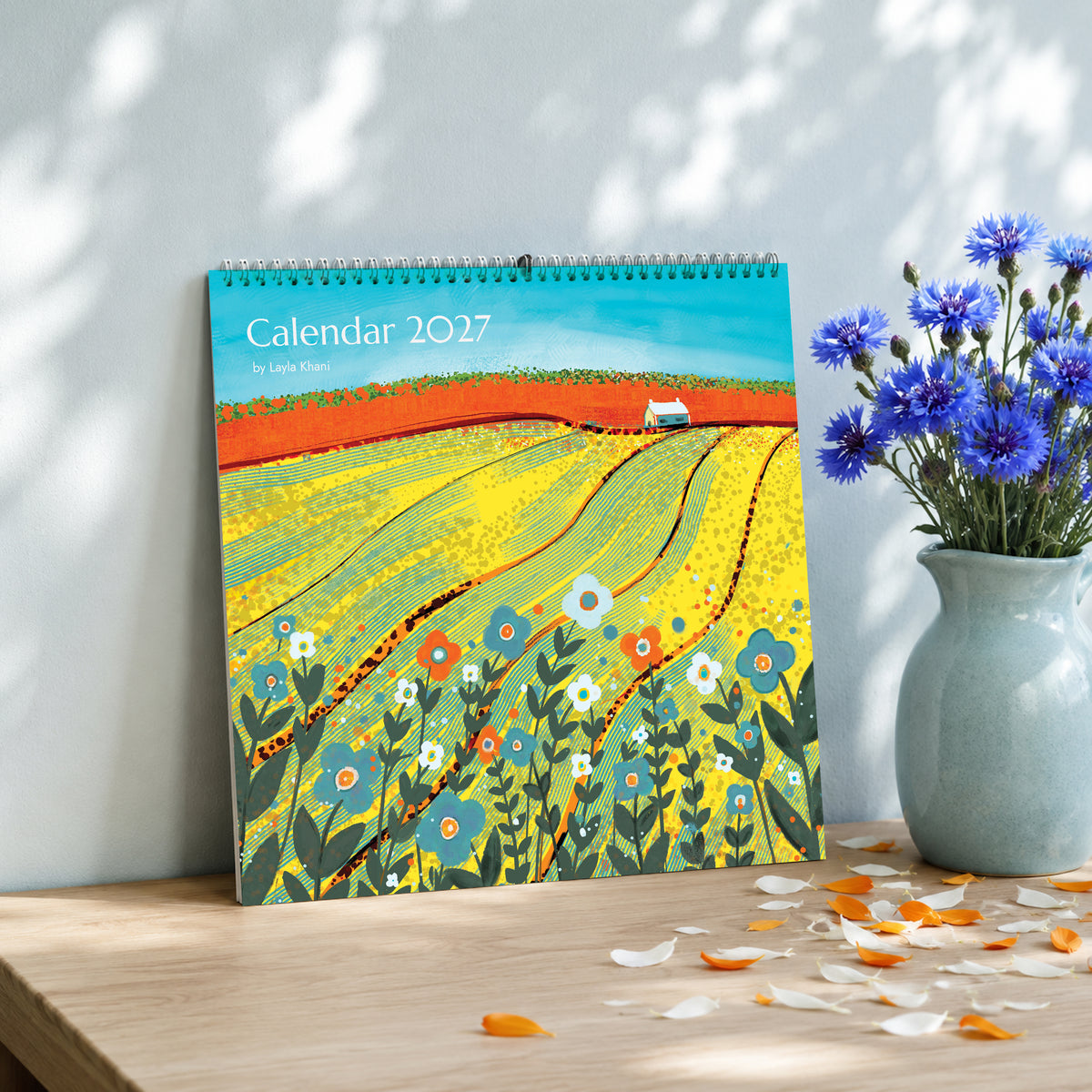 2027 large square art calendar with colourful countryside painting, displayed on a wooden table