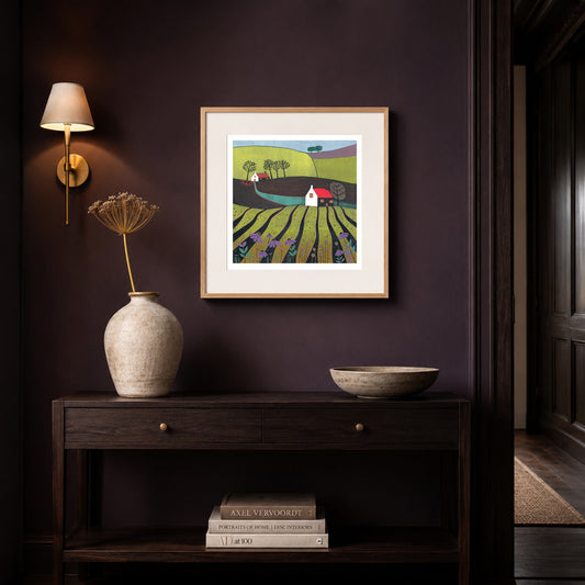 Linocut landscape print framed in oak on a deep aubergine wall above a dark console with a brass wall light.