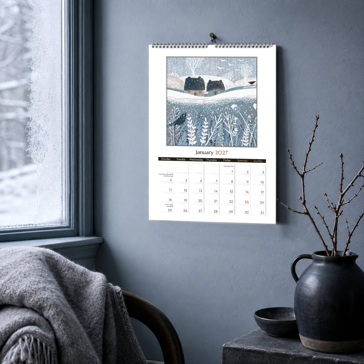 January 2027 wall calendar page featuring a snowy cottage landscape, displayed in a cool winter interior.