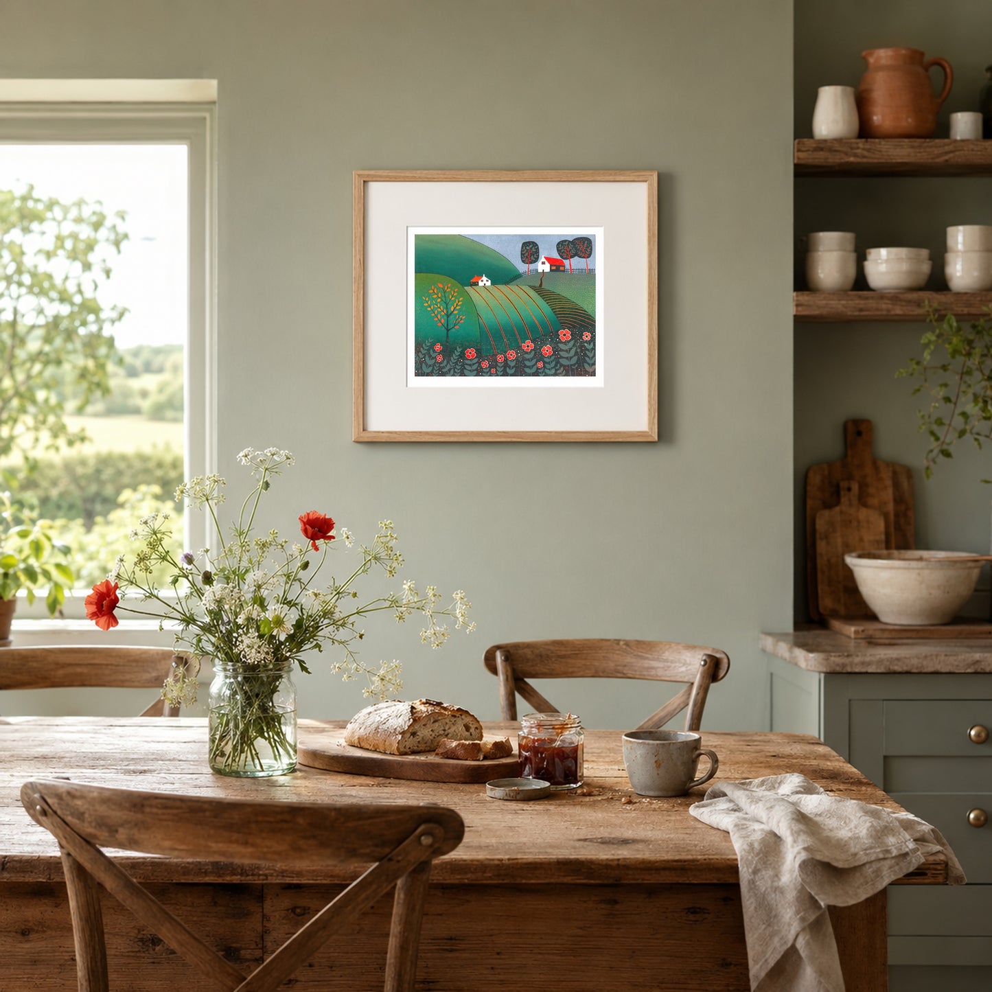 Framed linocut of a green poppy landscape on a sage wall beside a cream boucle armchair with a rust cushion and green throw.