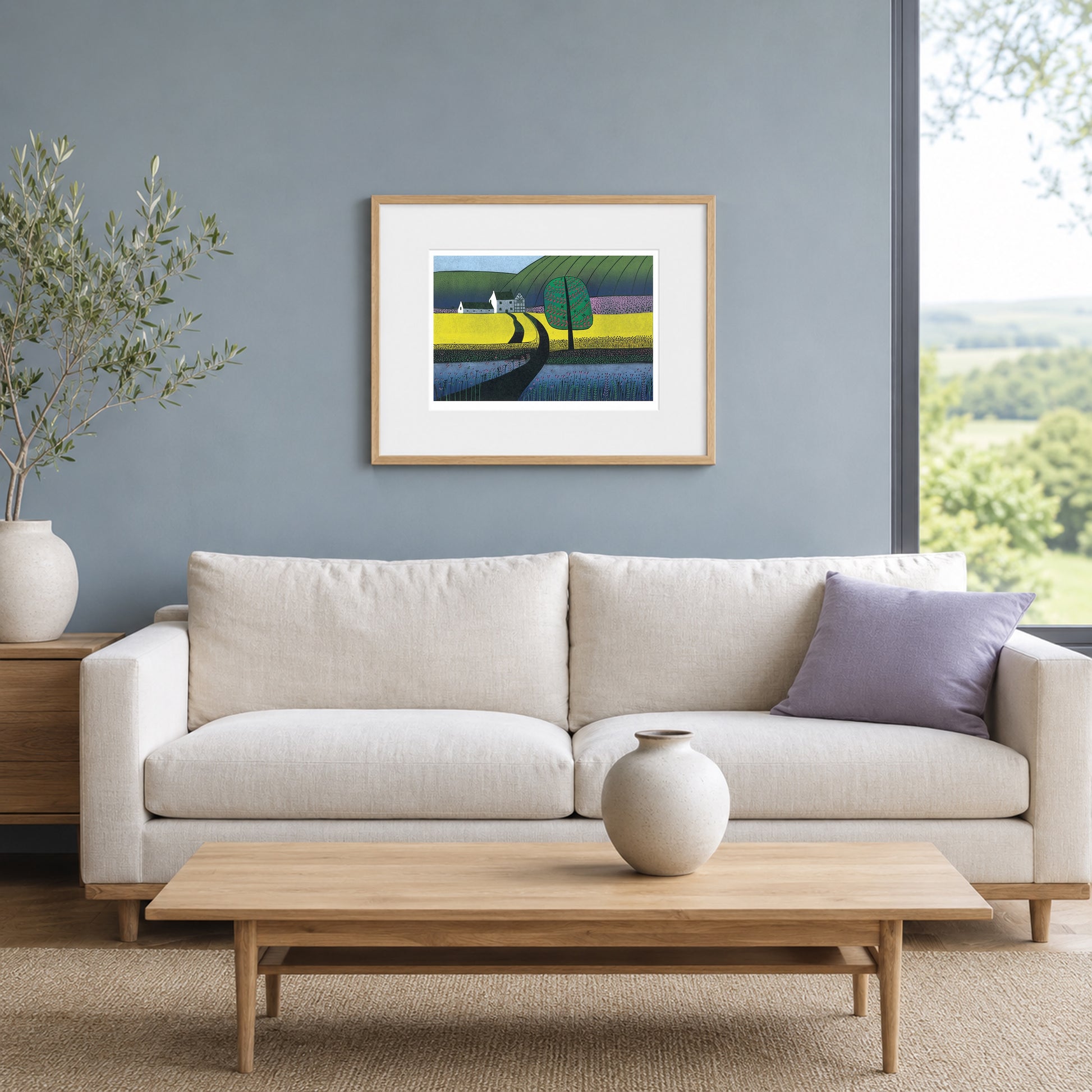 Framed linocut of a yellow and blue landscape on a slate-blue wall above a cream sofa with a lilac cushion.