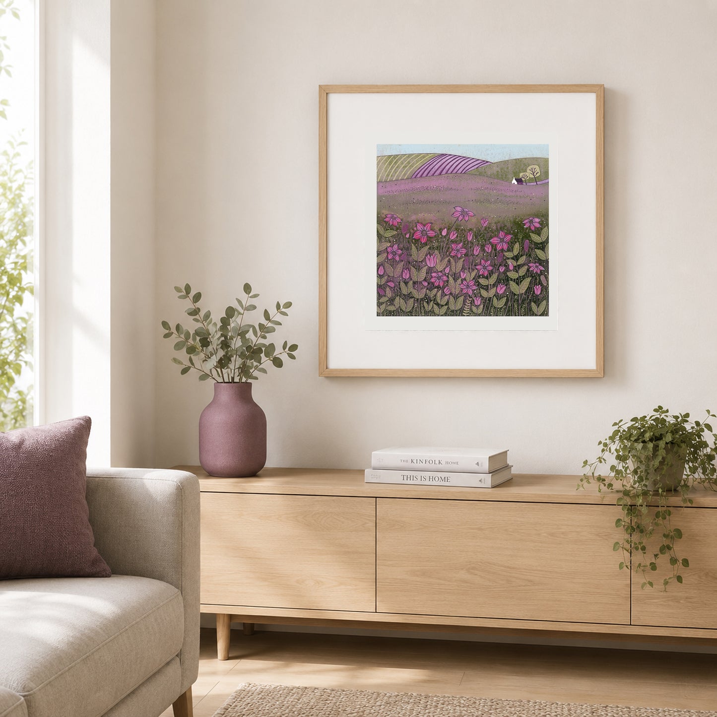Purple meadow linocut print in an oak frame above a pale wood console with a mauve vase and books.