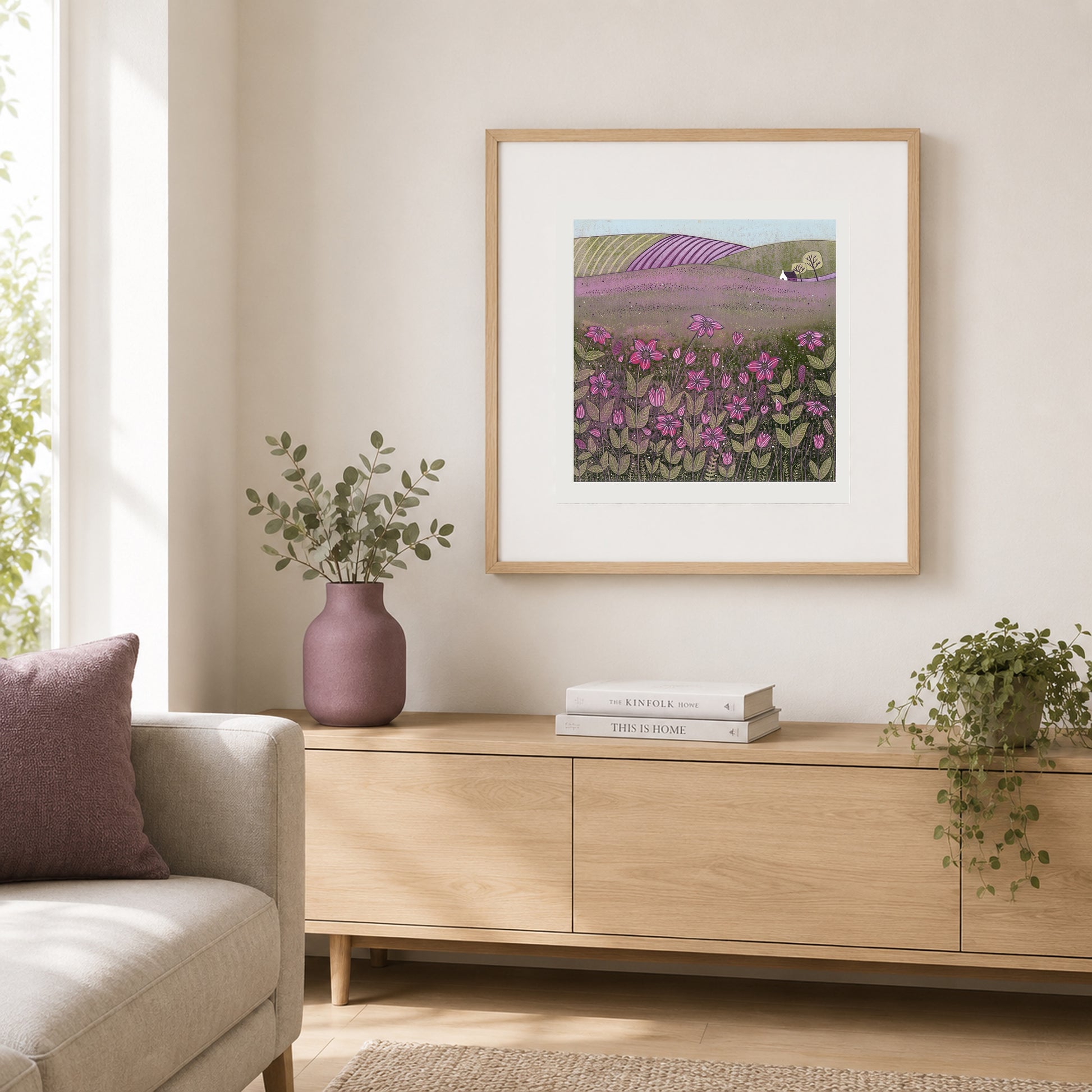 Purple meadow linocut print in an oak frame above a pale wood console with a mauve vase and books.