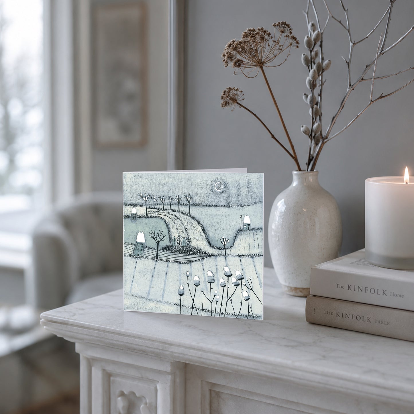 Blue-grey winter landscape Christmas card with trees, cottages and snow displayed on a mantel.