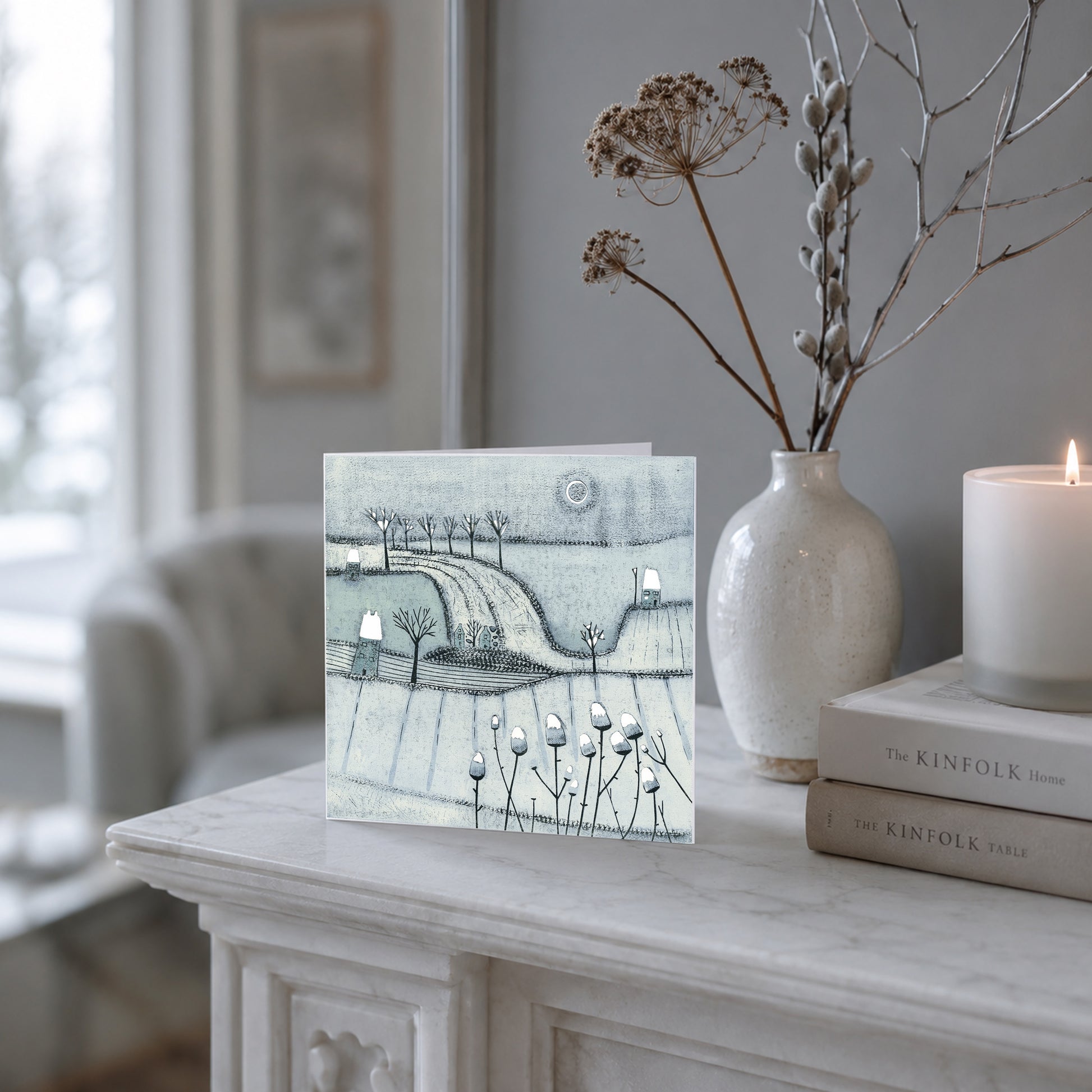 Blue-grey winter landscape Christmas card with trees, cottages and snow displayed on a mantel.