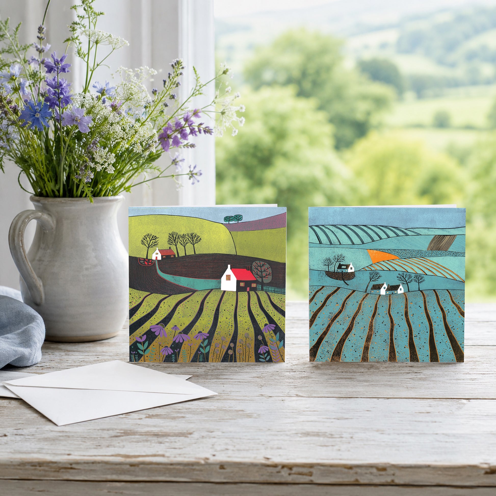Two linocut greeting cards of countryside scenes propped on a table with wildflowers in a stone jug