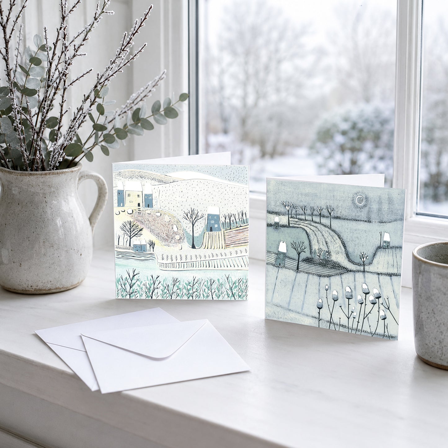 Two winter landscape Christmas cards reproduced from original linocut prints, displayed beside a snowy window.