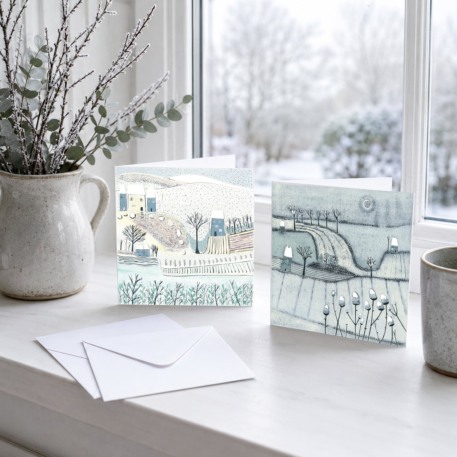 Two winter landscape Christmas cards reproduced from original linocut prints, displayed beside a snowy window.
