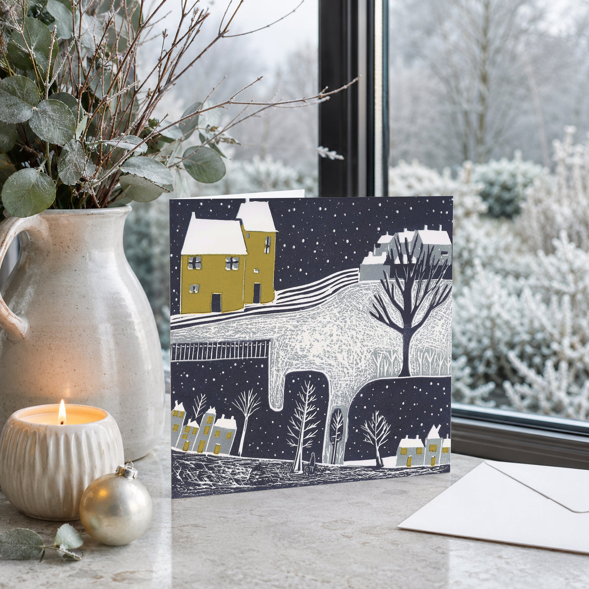 Wintry Nights Christmas card with a yellow house and snowy landscape displayed beside a winter window.