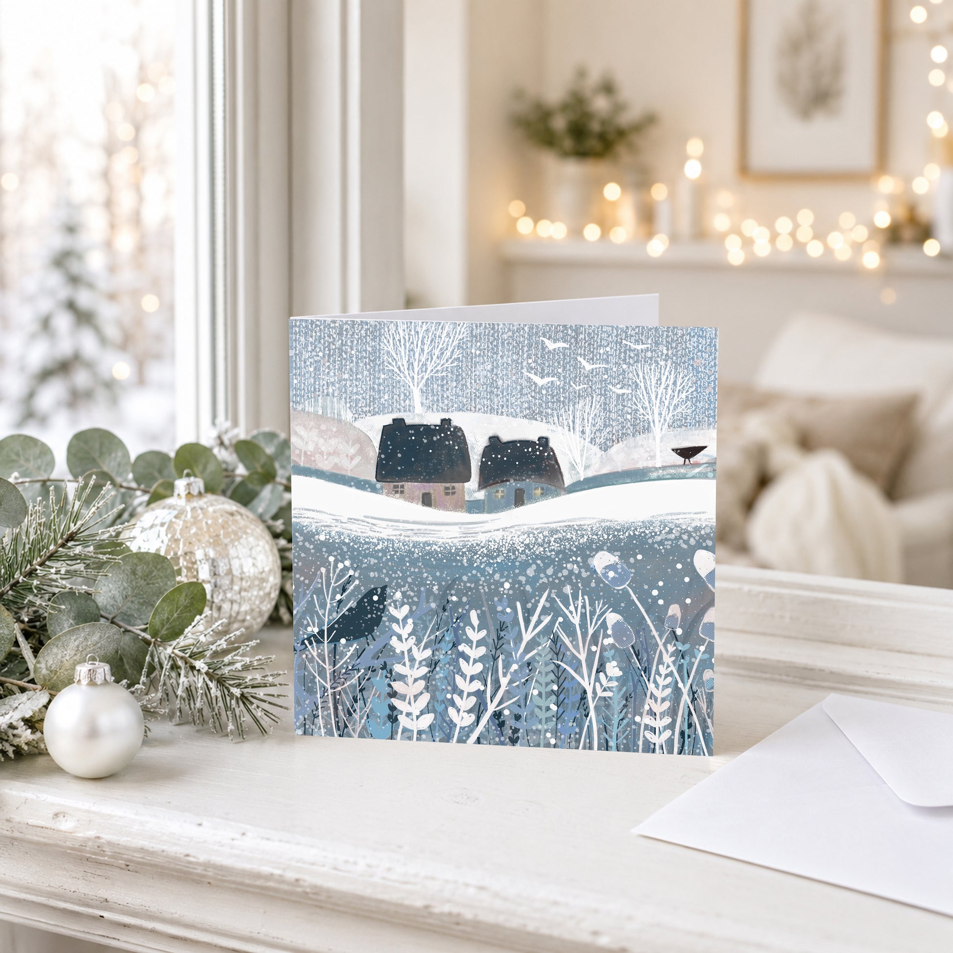 Winter landscape Christmas card featuring snowy cottages, winter plants and birds in soft blue and white