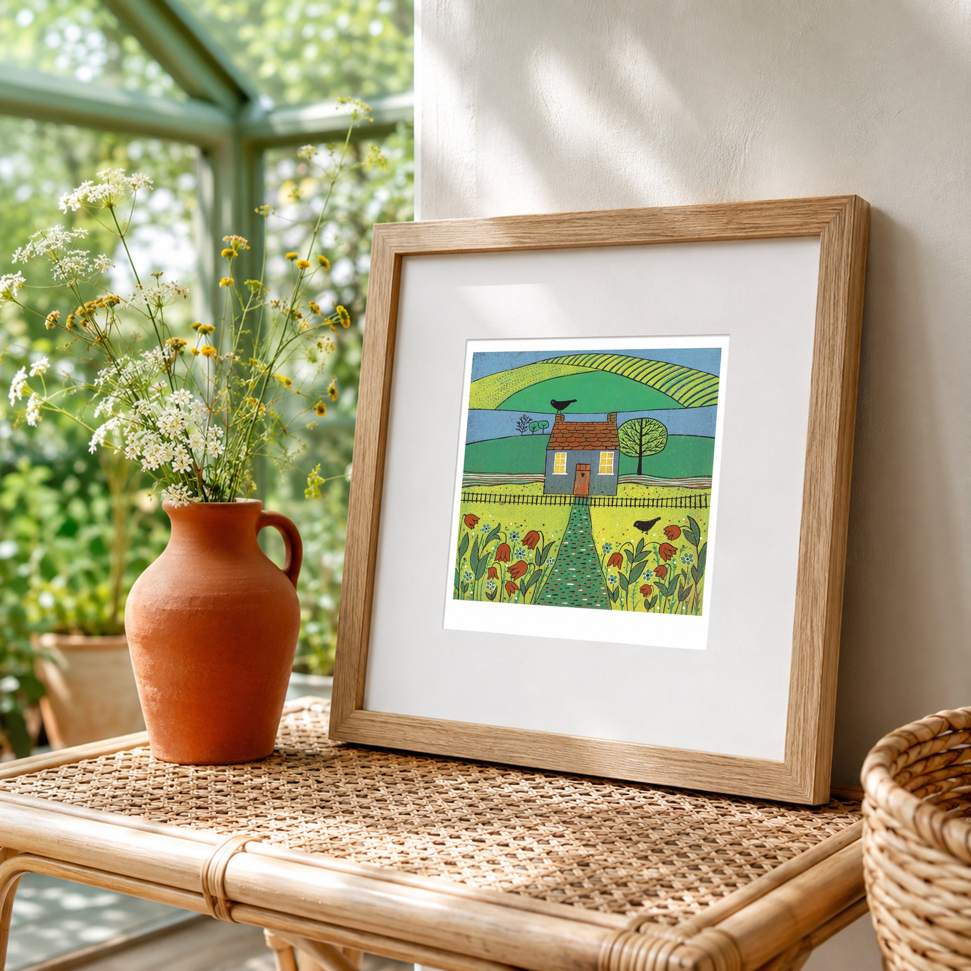 Framed Beyond the Garden linocut print with a cottage, birds and wildflowers displayed on a rattan table beside a sunlit window.